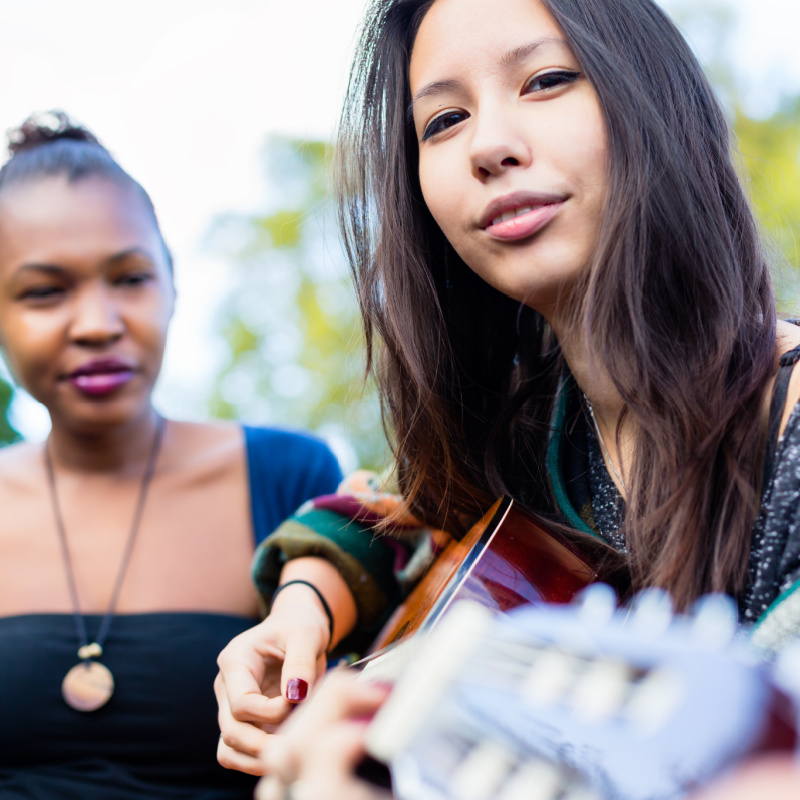 Asian young adult playing the acoustic guitar along with a Black friend.